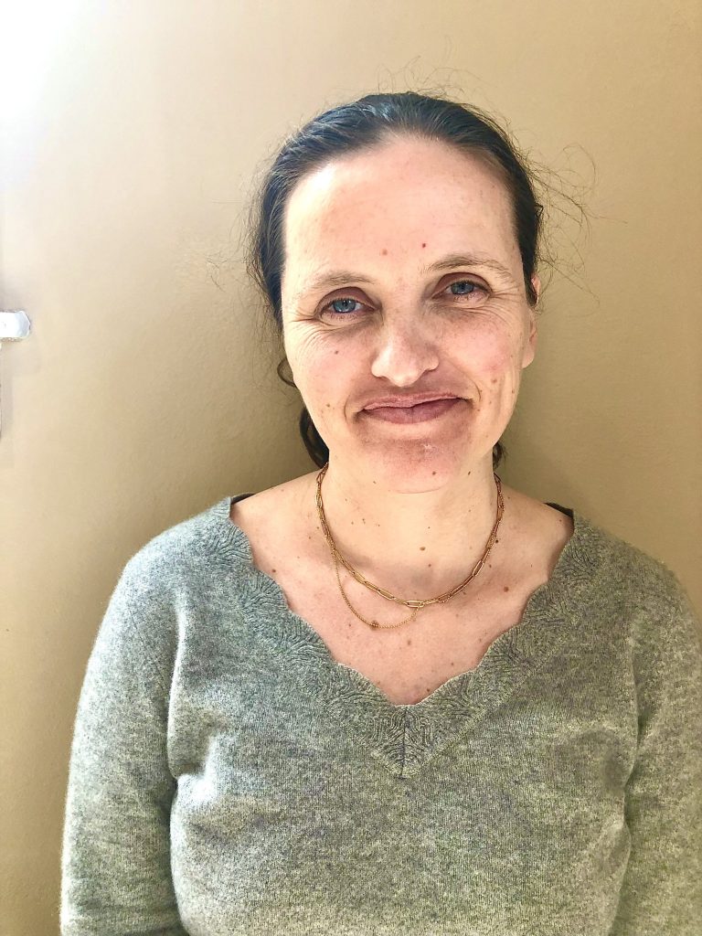 Photo featuring the top half of Alegria, front-facing. She is a white woman with brunette hair and she is sitting in front of a tan wall, smiling warmly. She is wearing a soft grey vneck sweater and a thin gold necklace.